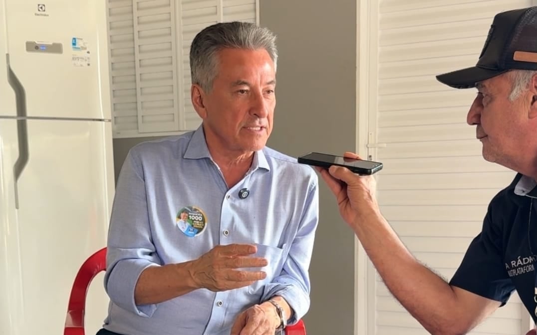 Roberto Hashioka, sentado, concede entrevista a um repórter de rádio que grava a conversa pelo celular.