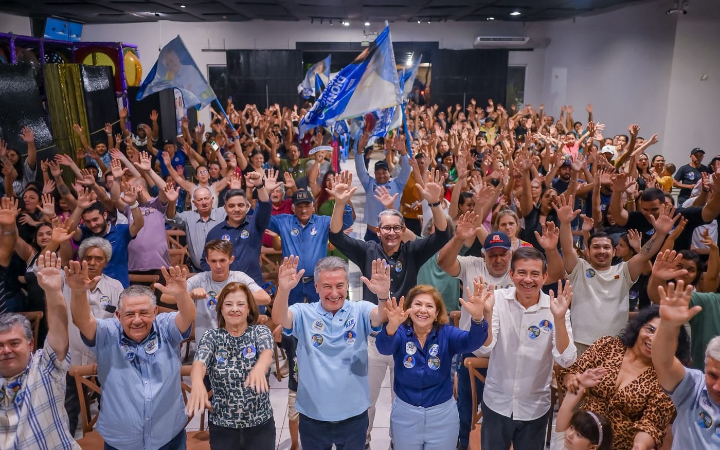 Salão lotado de apoiadores com as mãos erguidas e bandeiras azuis, com Roberto Hashioka e Dione Hashioka à frente, no lançamento das candidaturas em Dourados.