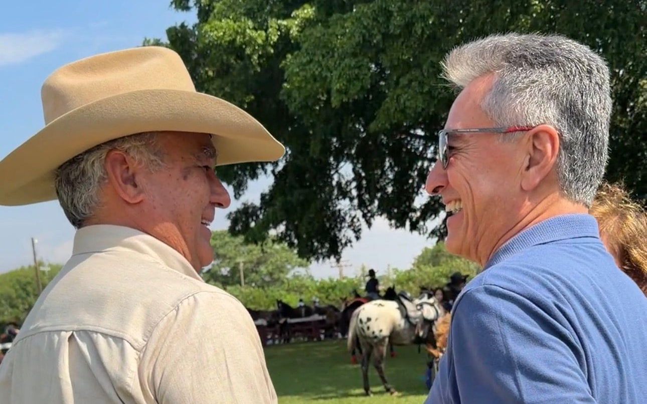 Roberto Hashioka conversa com um homem de chapéu de aba larga durante o desfile de cavaleiros, com cavalos e cavaleiros ao fundo.