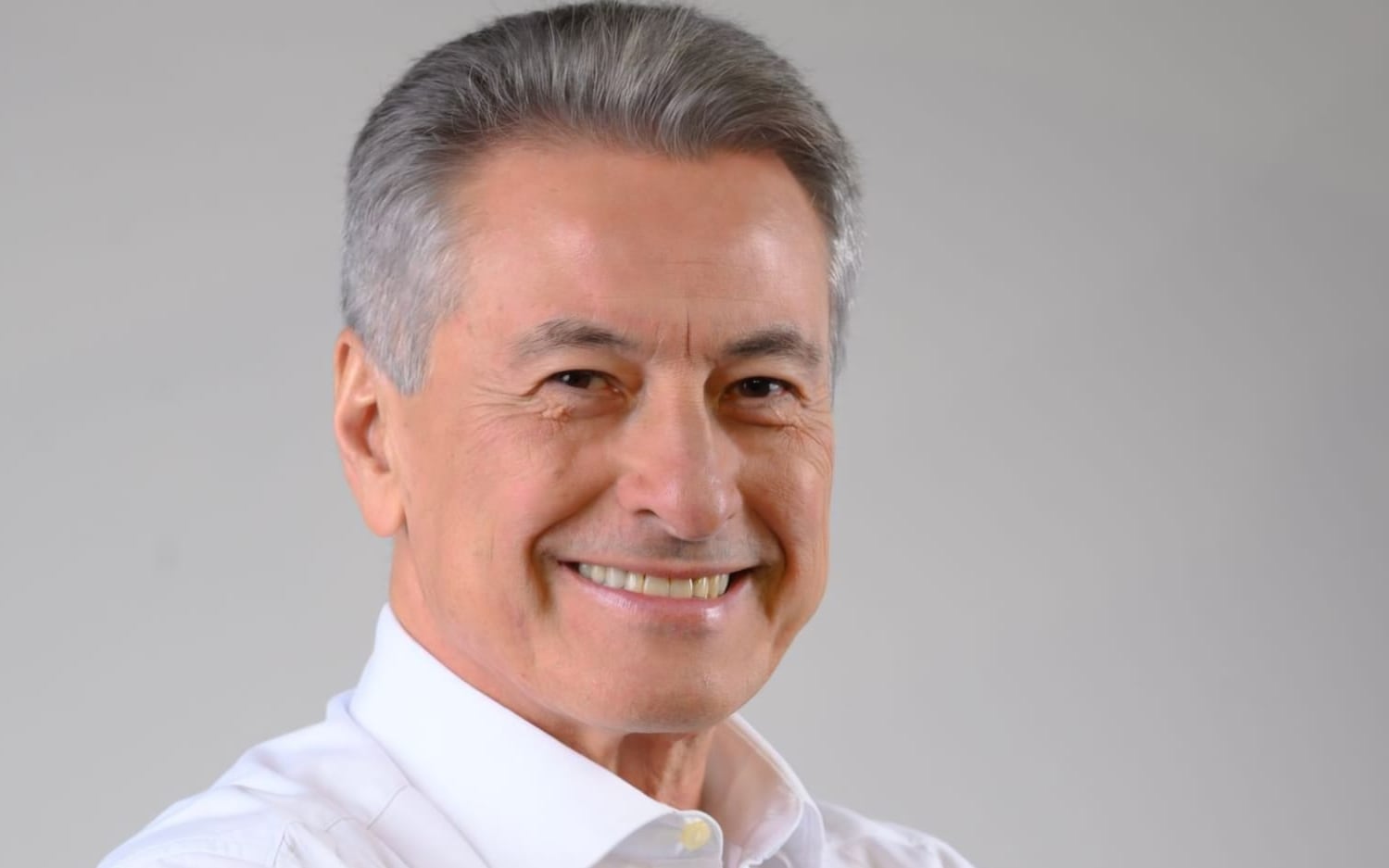 Retrato de estúdio de Roberto Hashioka, sorrindo, camisa branca, fundo cinza.