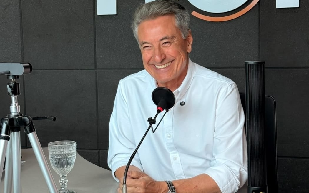 Roberto Hashioka, de camisa branca, sorri sentado diante do microfone no estúdio da Rádio Hora, em Campo Grande.