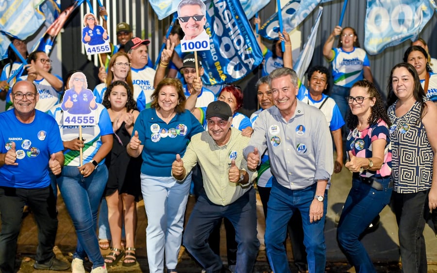 Roberto Hashioka sorri ao lado de apoiadores durante caminhada de campanha à noite, cercado por bandeiras e cartazes com seu número.