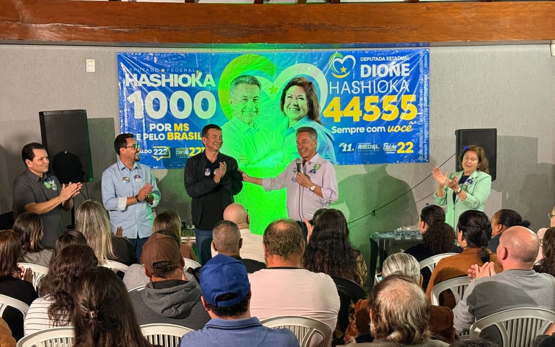 Roberto Hashioka aperta a mão de um aliado no palco, sob um banner de campanha, diante de uma plateia sentada.